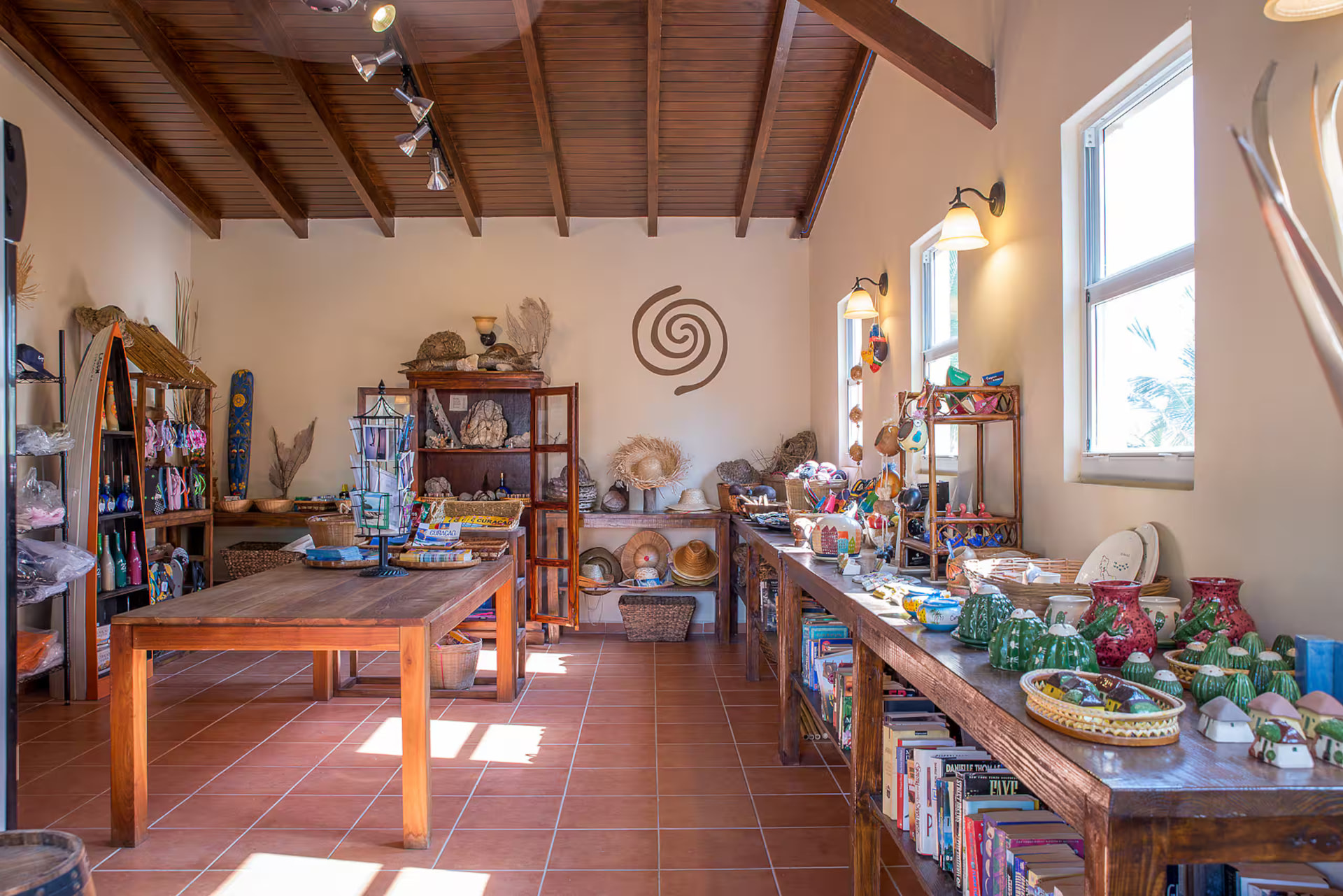 Interior of the resort's shop: wooden shelves filled with books, ceramics and straw hats, a table with postcards and guides, terracotta floor and a dark beamed ceiling