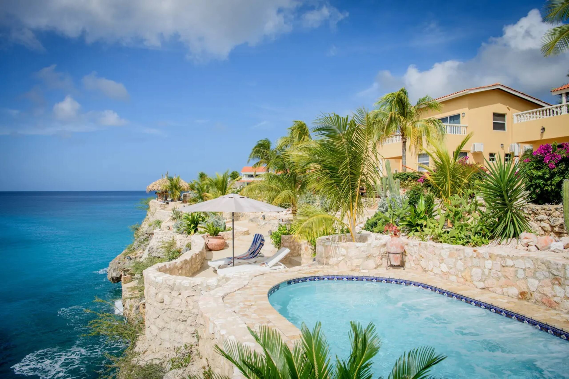 Cliffside terrace with an oval plunge pool, sun loungers and an open umbrella beside a peach-coloured building with bougainvillea, the open sea beyond